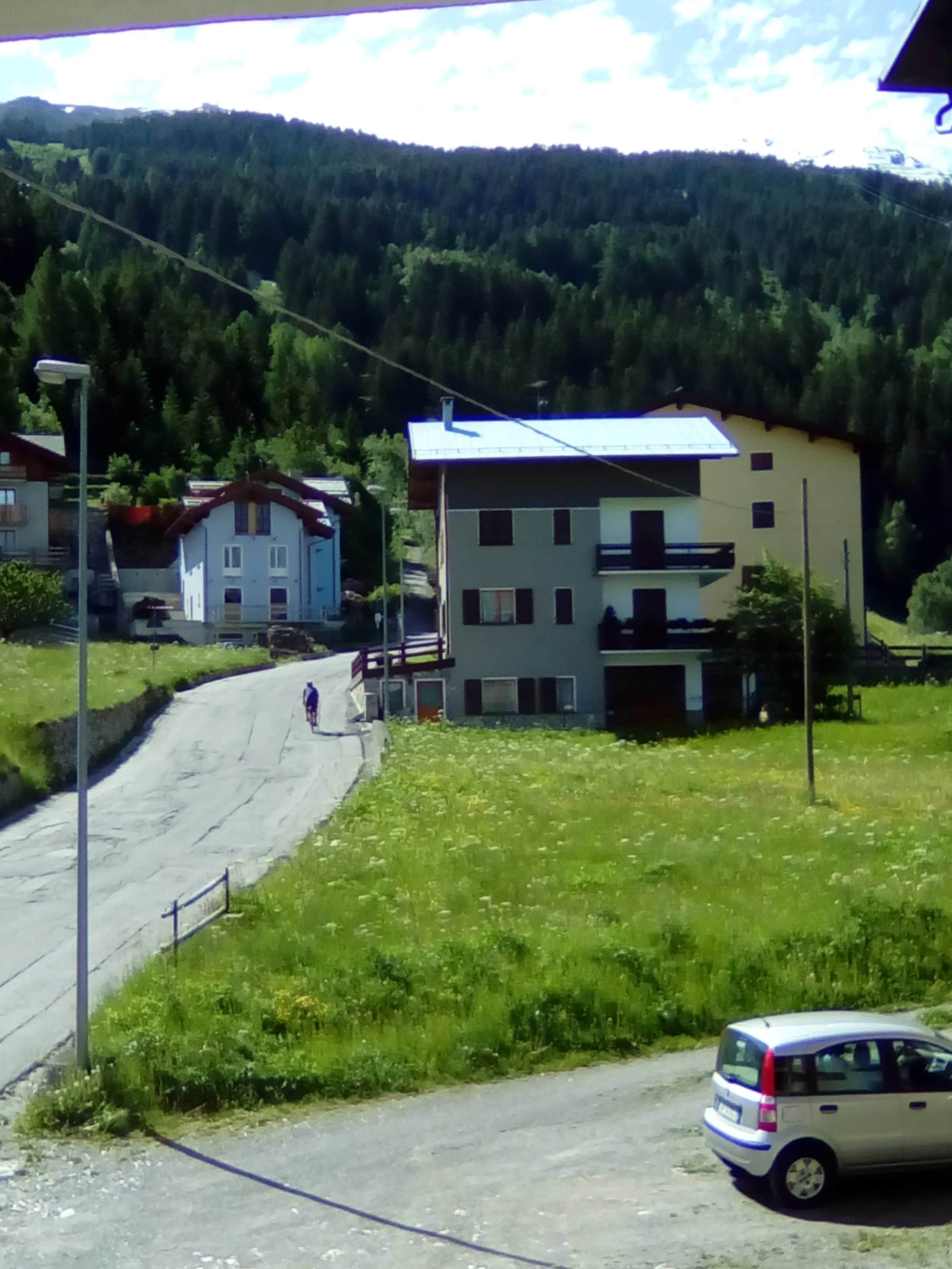 A road curving through a rural area with houses on one side and a forested hill in the background.