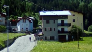 Une route sinueuse traverse une zone rurale, avec des maisons d’un côté et une colline boisée en arrière-plan.
