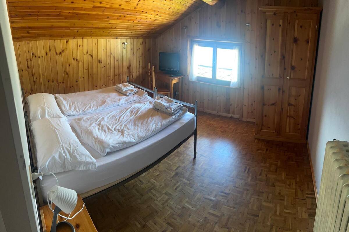 Una habitación con paredes y suelo de madera, una cama con ropa blanca, un televisor sobre una mesa de madera y una ventana que deja entrar luz natural.