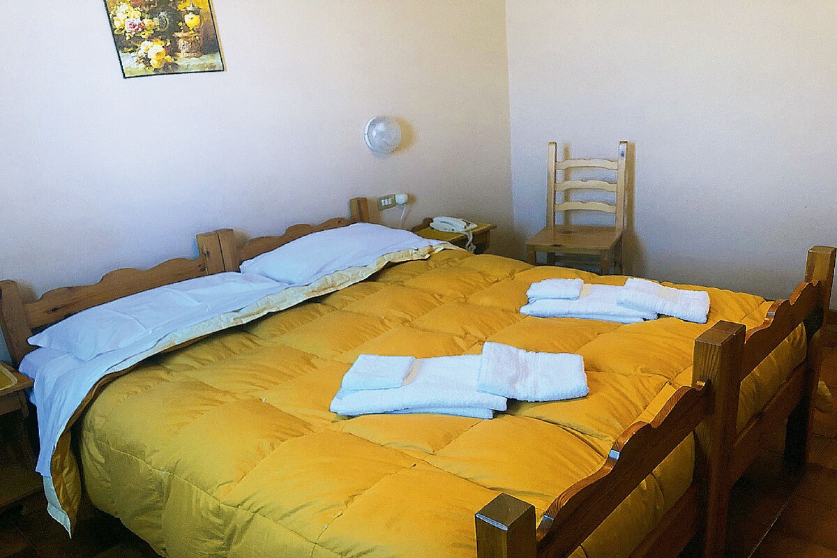 A double bed with a yellow bedspread, two white towels, a wooden headboard, a bedside table with a lamp, and a chair.