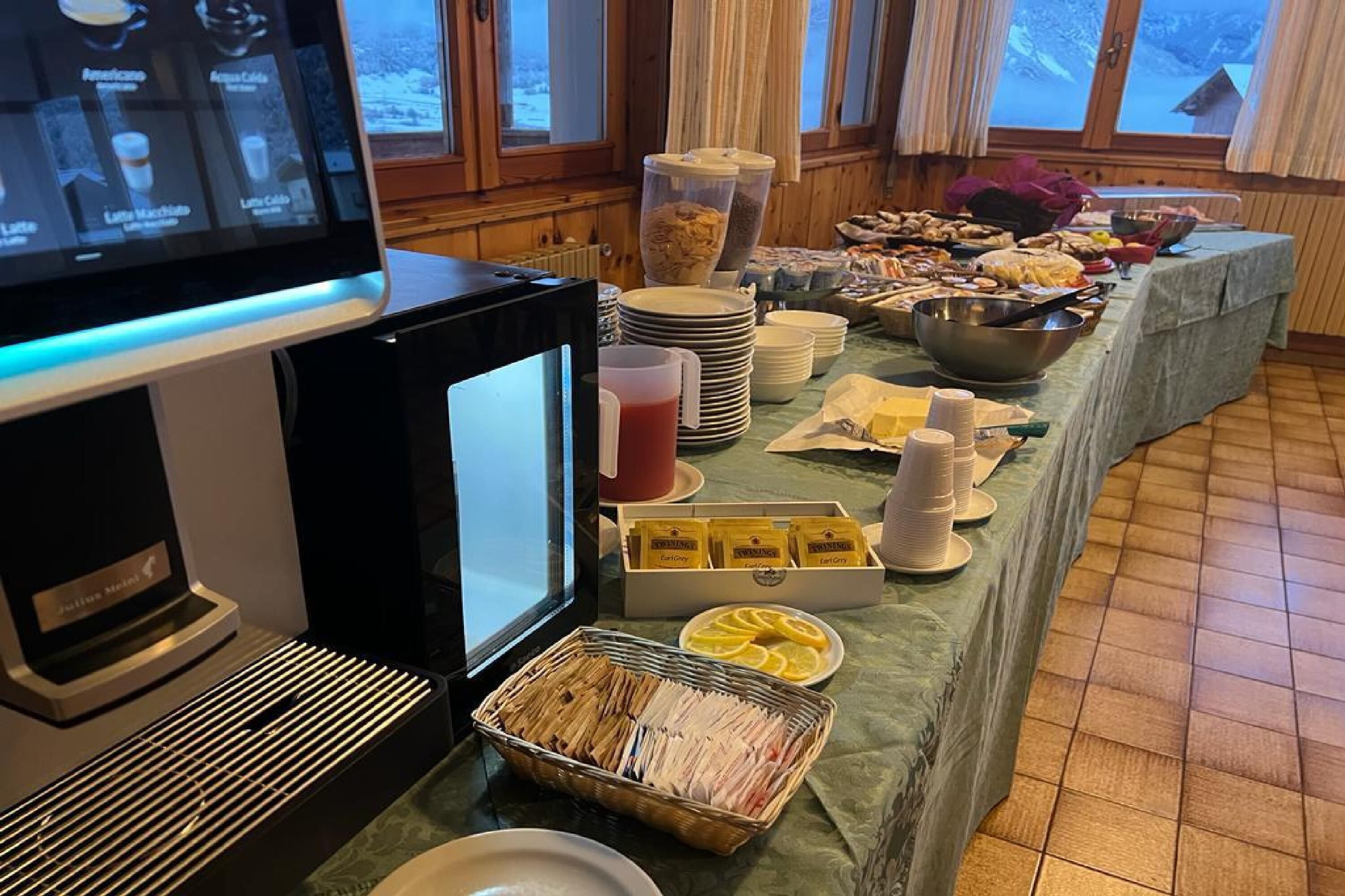 A buffet setup with various food items, a beverage station, and a view of the outdoors through large windows.