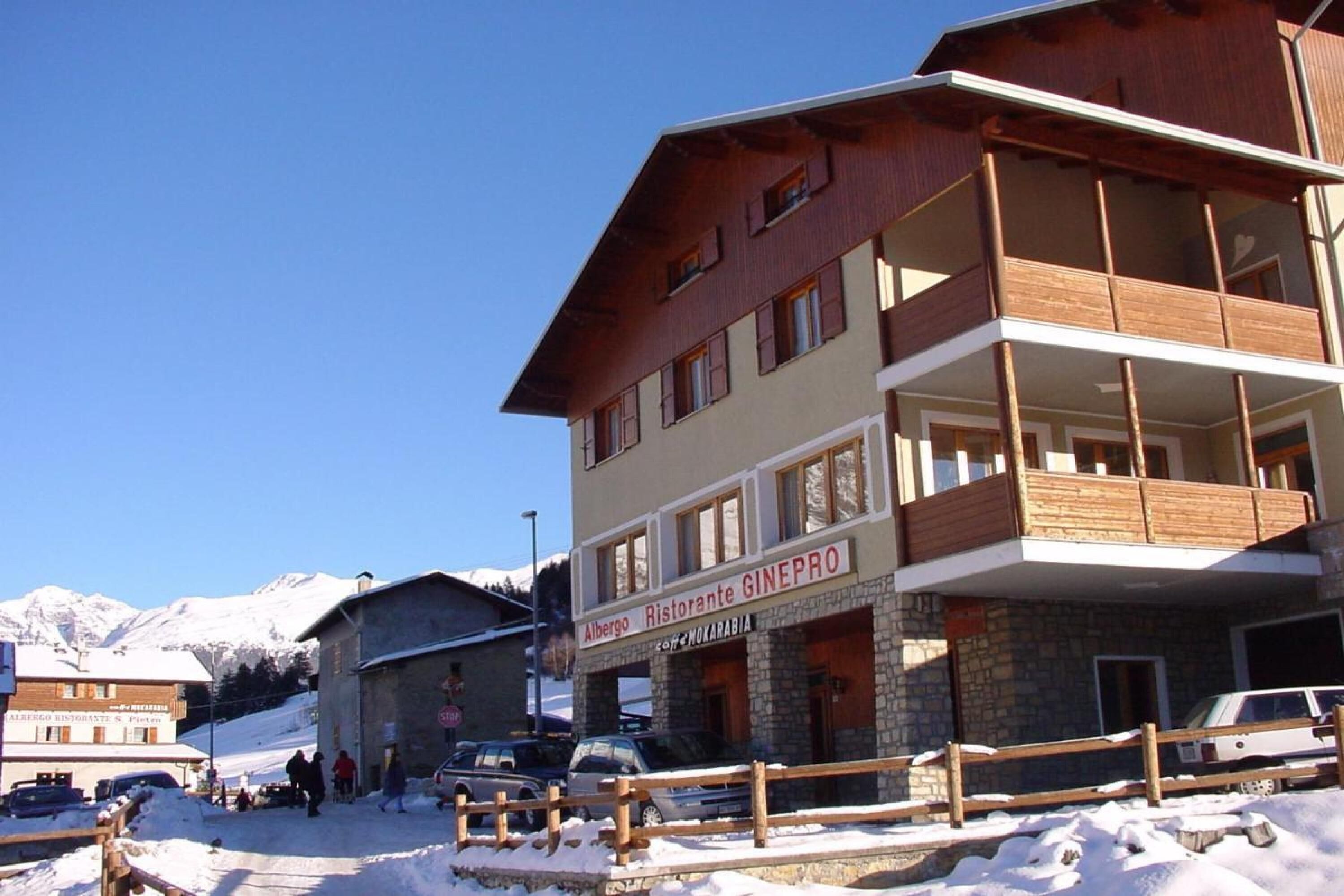 A multi-story building with a restaurant named "Albergo Ristorante GINEPRO" on the ground floor. There are several cars parked in front, and a wooden fence separates the building from the snow-covered ground.