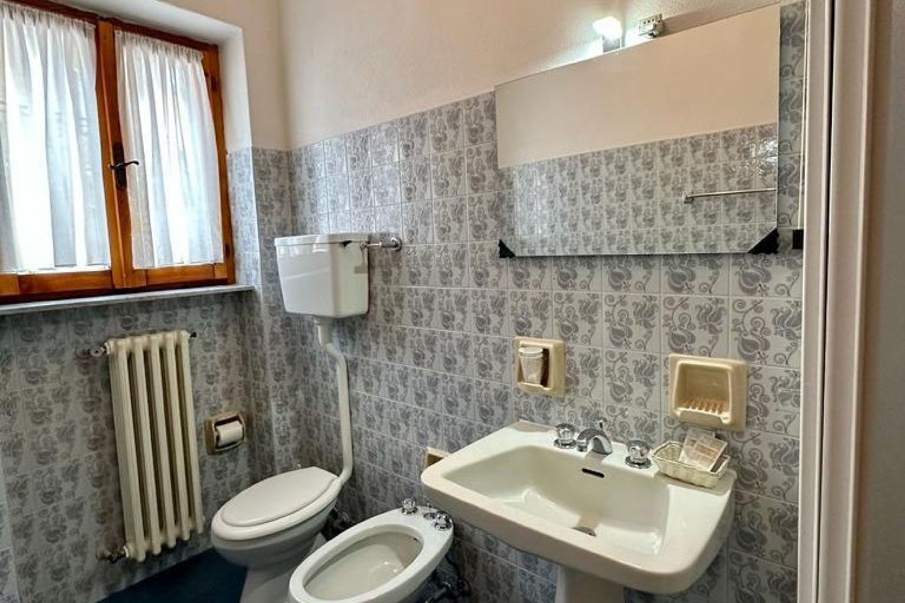 Un bagno con toilette, bidet, lavandino e una finestra con tende.