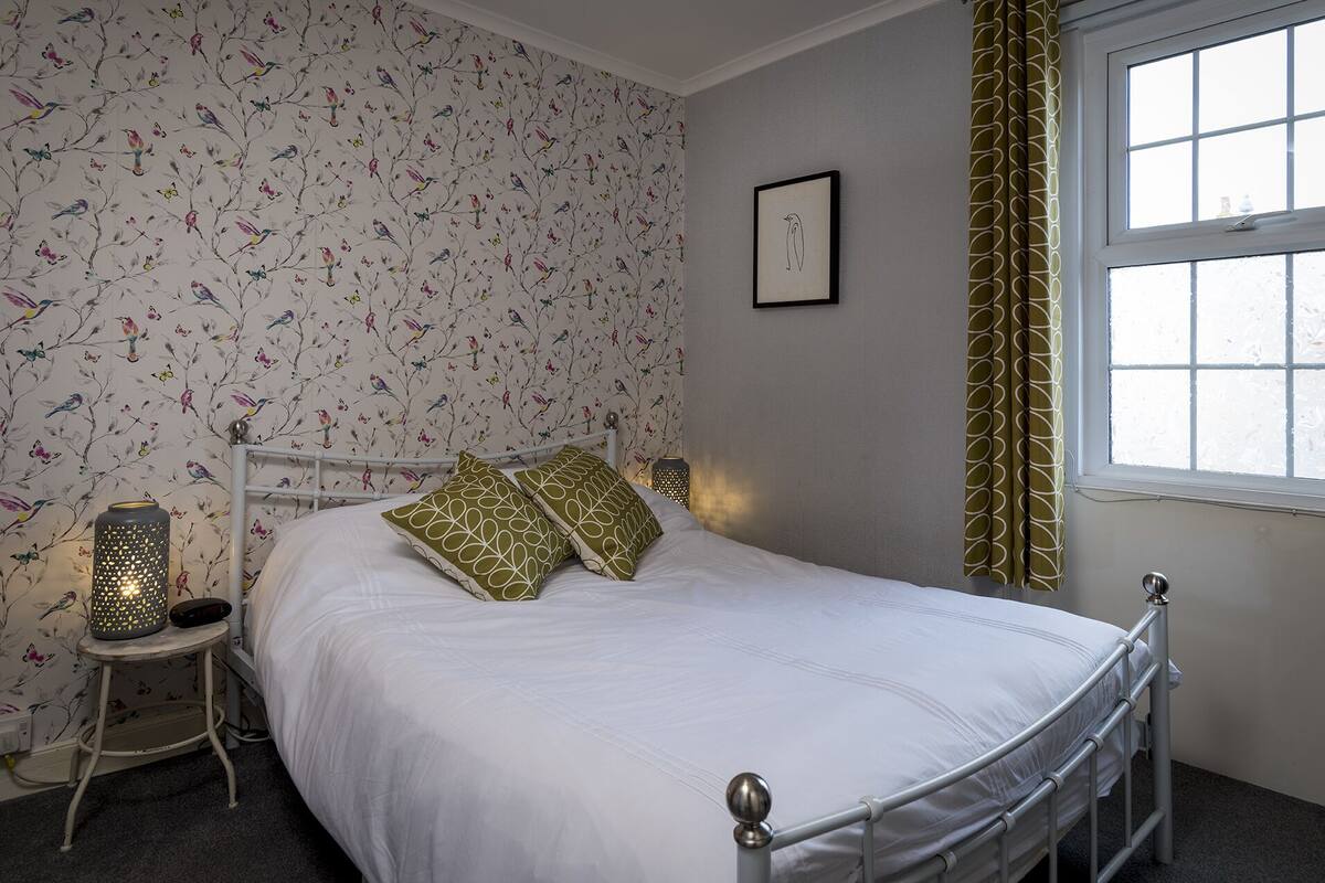 Ein Schlafzimmer mit einem Bett, einem Nachttisch, einer Wandlampe, einem Fenster mit Vorhängen und einer Wand mit Vogelmuster.
