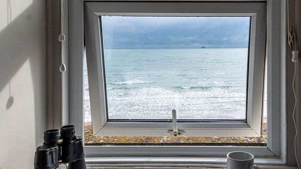 A window with binoculars and a mug on the windowsill, overlooking a beach with waves and a cloudy sky. - Channel View Guest House (Weymouth)