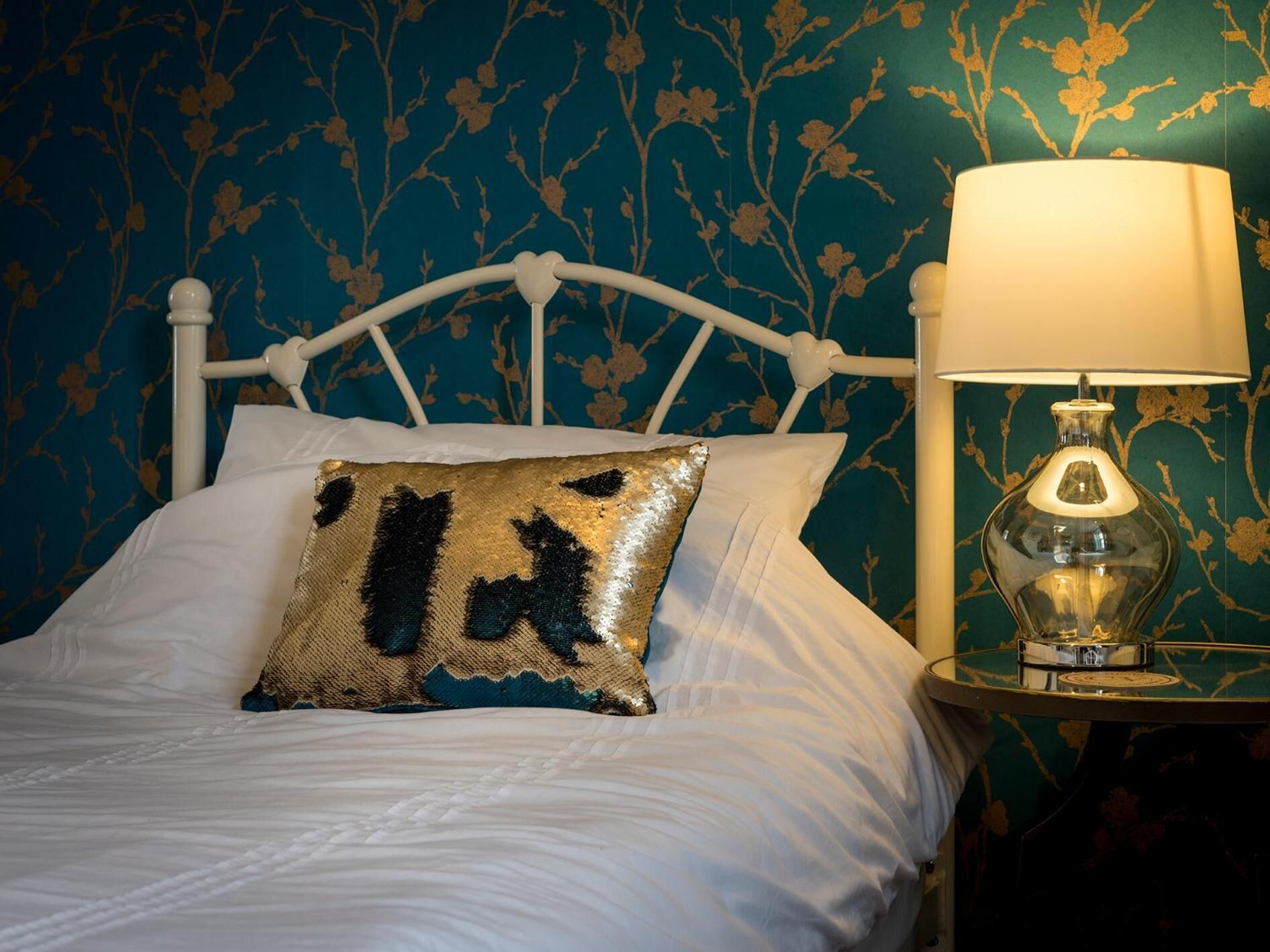 A bed with white bedding and a decorative pillow, a white metal headboard, and a bedside table with a lamp.