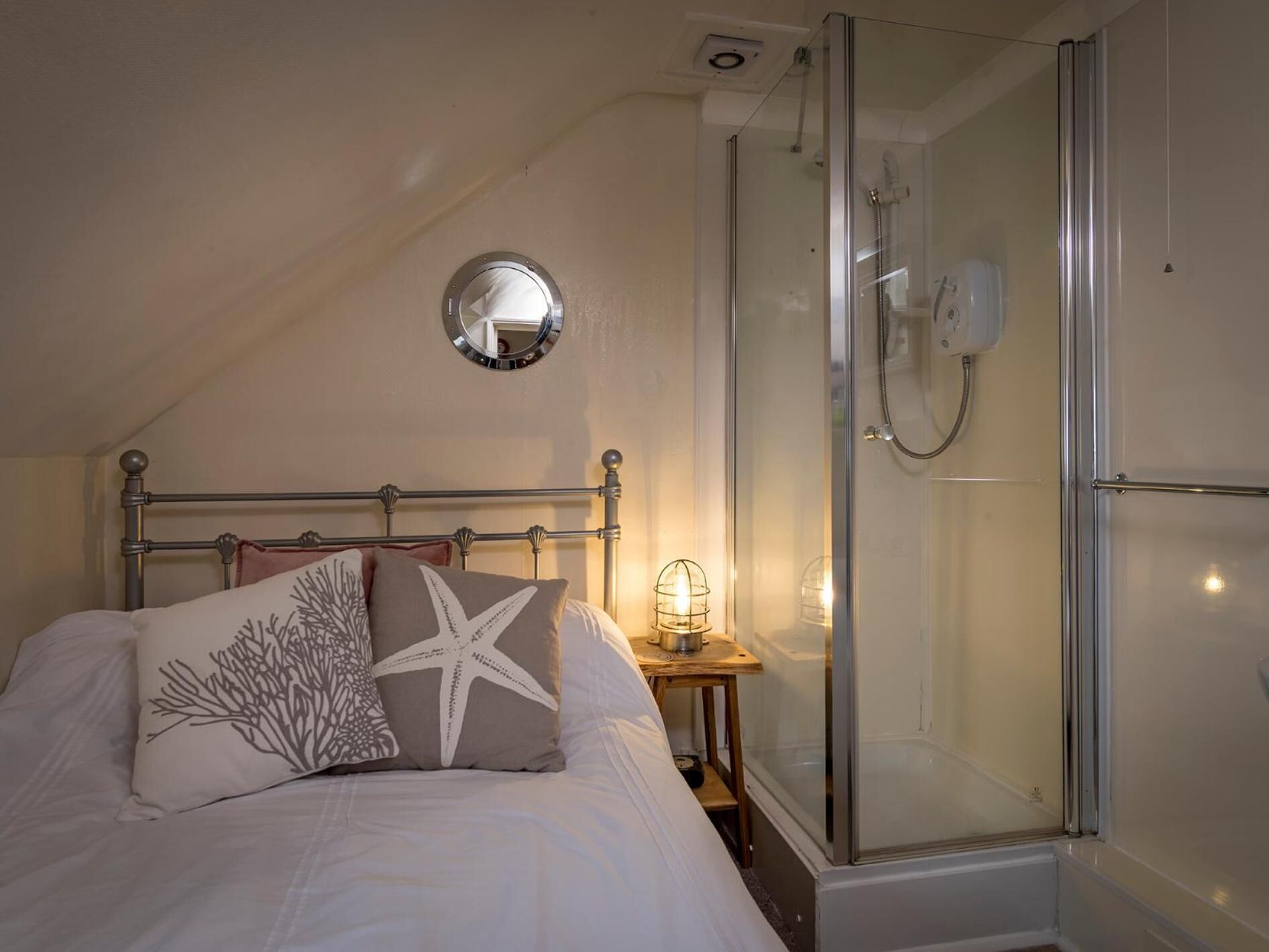 A bedroom with a bed, a round mirror, a nightstand with a lamp, and a glass-enclosed shower.