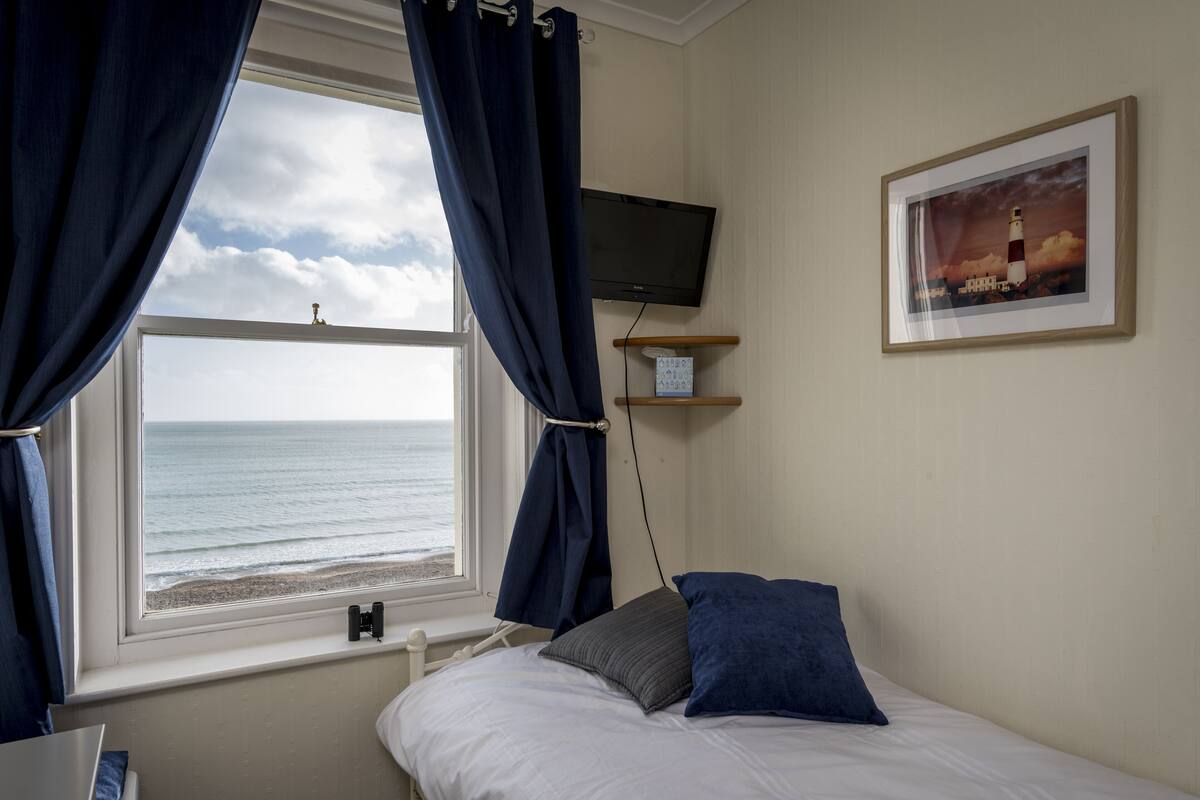 Ein Schlafzimmer mit Bett, Fernseher, Fenster mit Vorhängen, Ablagefläche mit Taschentuchbox und einem gerahmten Bild an der Wand.
