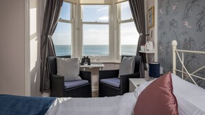 A room with a bed, two wicker chairs, a small table with a glass, and a view of the sea through a bay window. - Channel View Guest House (Weymouth)