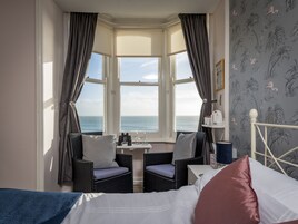 Una habitación con una cama, dos sillas de mimbre, una mesita con un vaso y vistas al mar a través de un ventanal.
