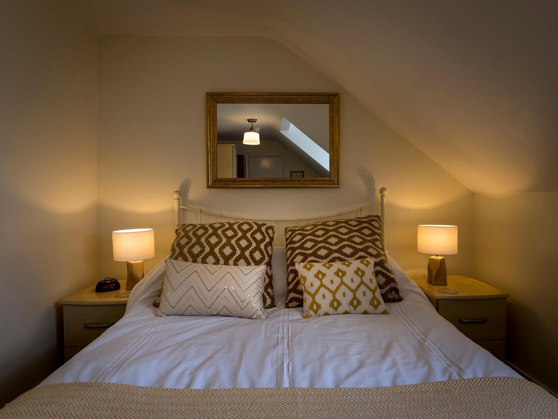 Un dormitorio con una cama, dos mesitas de noche con lámparas, un espejo sobre la cama y almohadas estampadas.