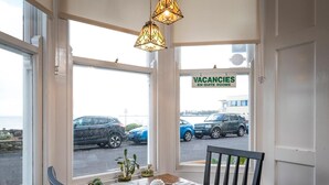 A dining area with a table set for four, a view of the street with parked cars, and a sign advertising vacancies in en-suite rooms. - Channel View Guest House (Weymouth)