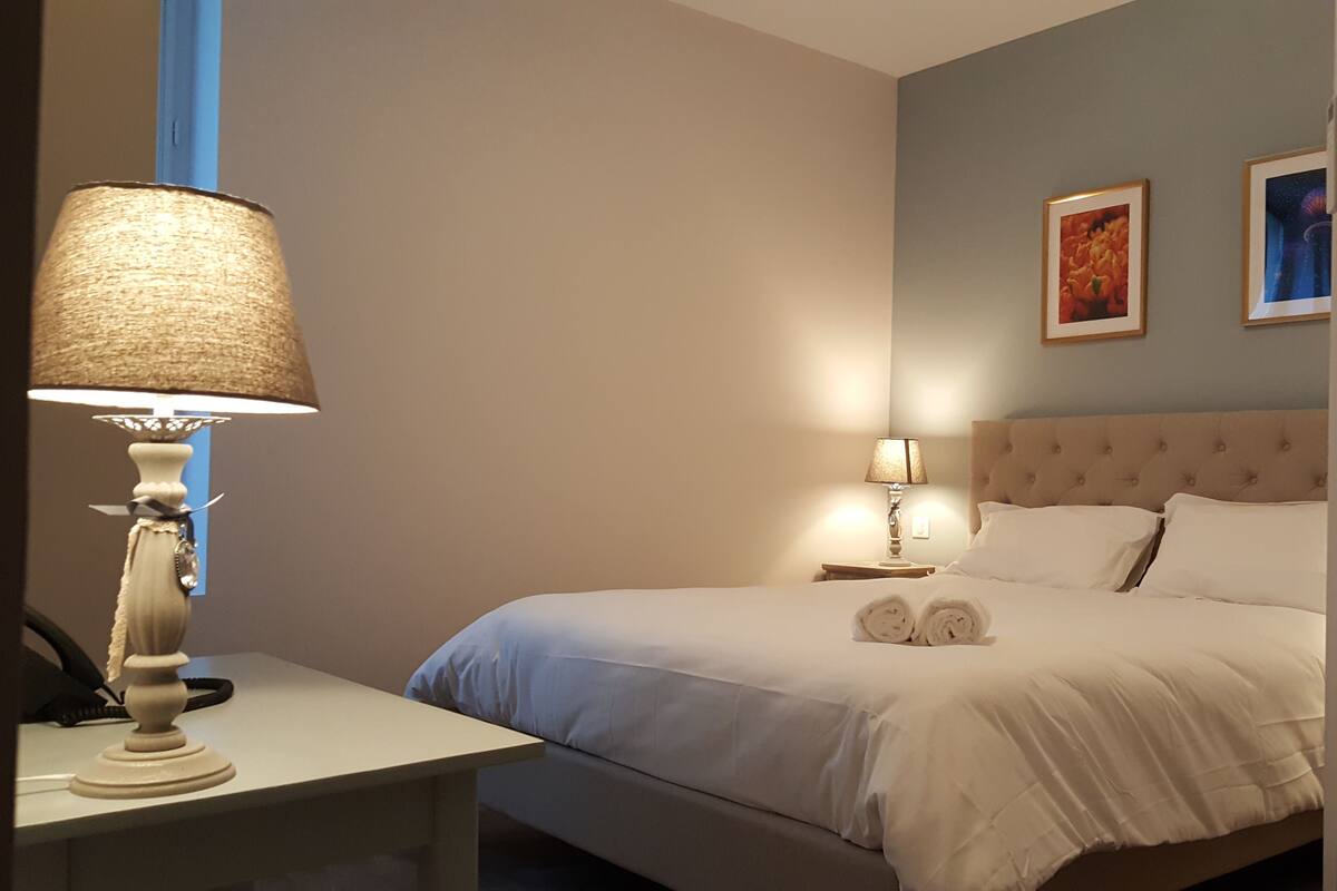 A neatly made bed with white linens, a bedside lamp, and a nightstand with a phone. There are two framed pictures on the wall.