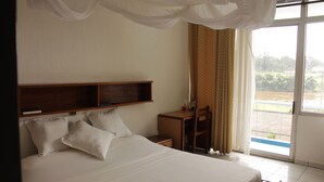 A hotel room with a large bed, a wooden headboard, a desk, and a chair. There is a window with curtains and a view of greenery outside. - Hostellerie De La Sanaga (Edea)