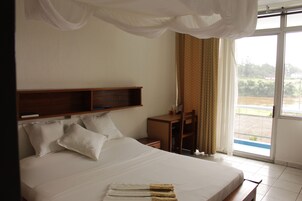 Quarto de hotel com uma cama grande, cabeceira de madeira, escrivaninha e uma cadeira. Há uma janela com cortinas e vista para o verde lá fora.