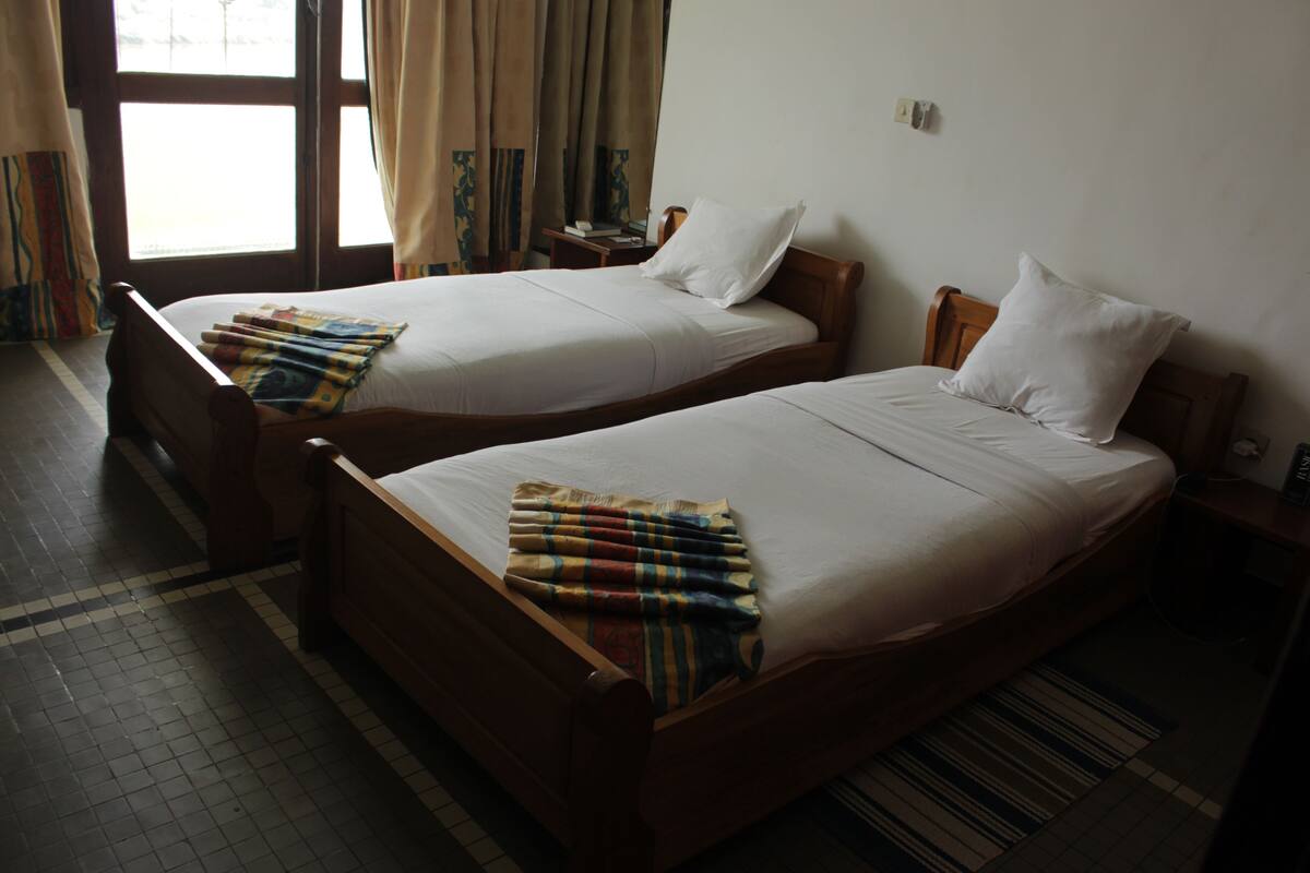 Two single beds with wooden frames and white linens, a small bedside table with a book and a lamp, and a window with curtains.