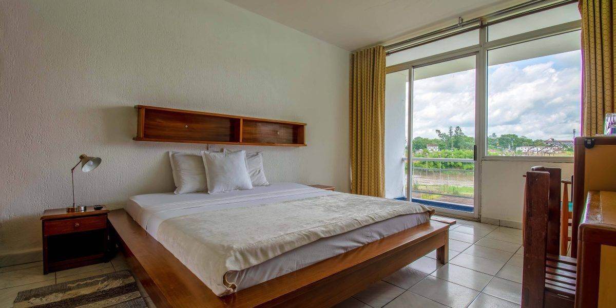 Quarto com cama grande, mesa de cabeceira, abajur e vista para o exterior através de uma janela ampla.
