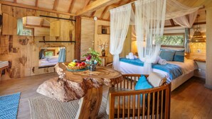 Une chambre dans une cabane en bois, avec un lit à baldaquin, une table à manger en bois sur laquelle est posé un panier de fruits, et une chaise rustique.