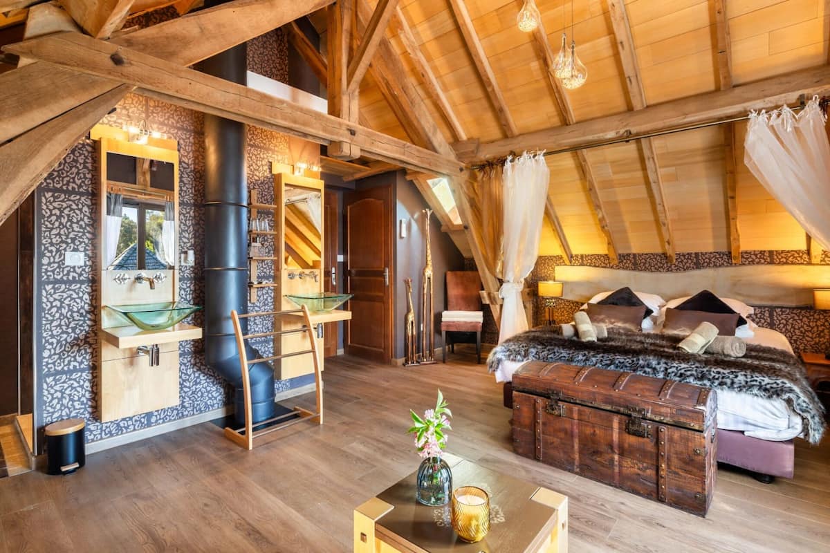 Een slaapkamer op zolder met hoge plafonds, zichtbare houten balken, een wastafel met glazen deurtjes, een bed met een bontdeken en een vintage koffer.