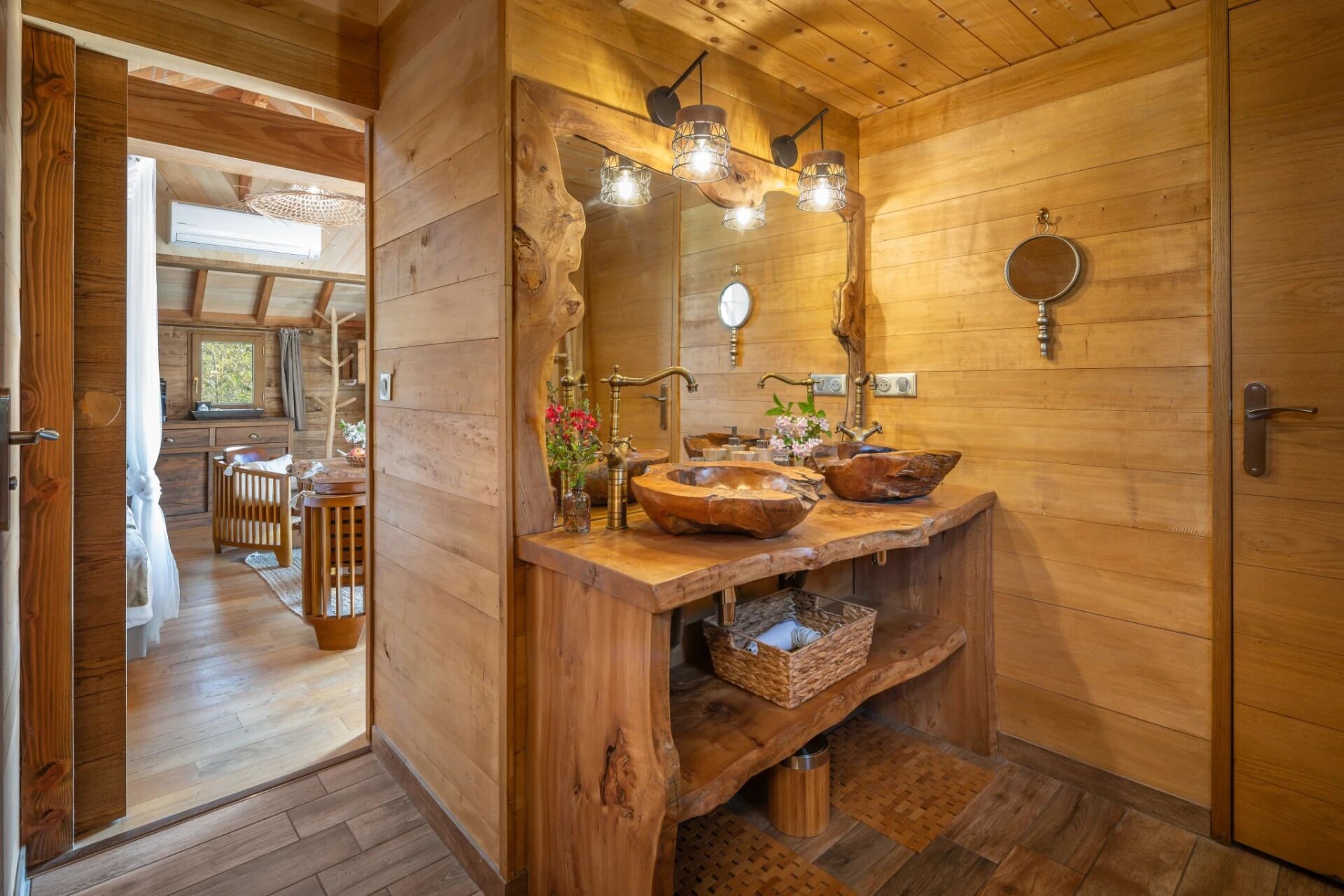 Une salle de bain rustique en bois, dotée d’un grand miroir, de deux éviers en forme de bols en bois et d’une étagère en bois sur laquelle repose un panier.
