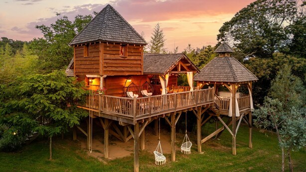 Une cabane en bois dotée d’une terrasse et de deux petits pavillons, le tout entouré d’une végétation luxuriante.