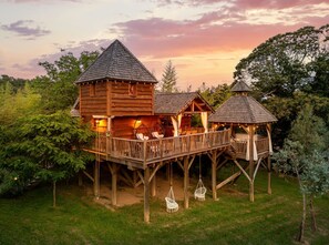 Une cabane en bois dotée d’une terrasse et de deux petits pavillons, le tout entouré d’une végétation luxuriante.