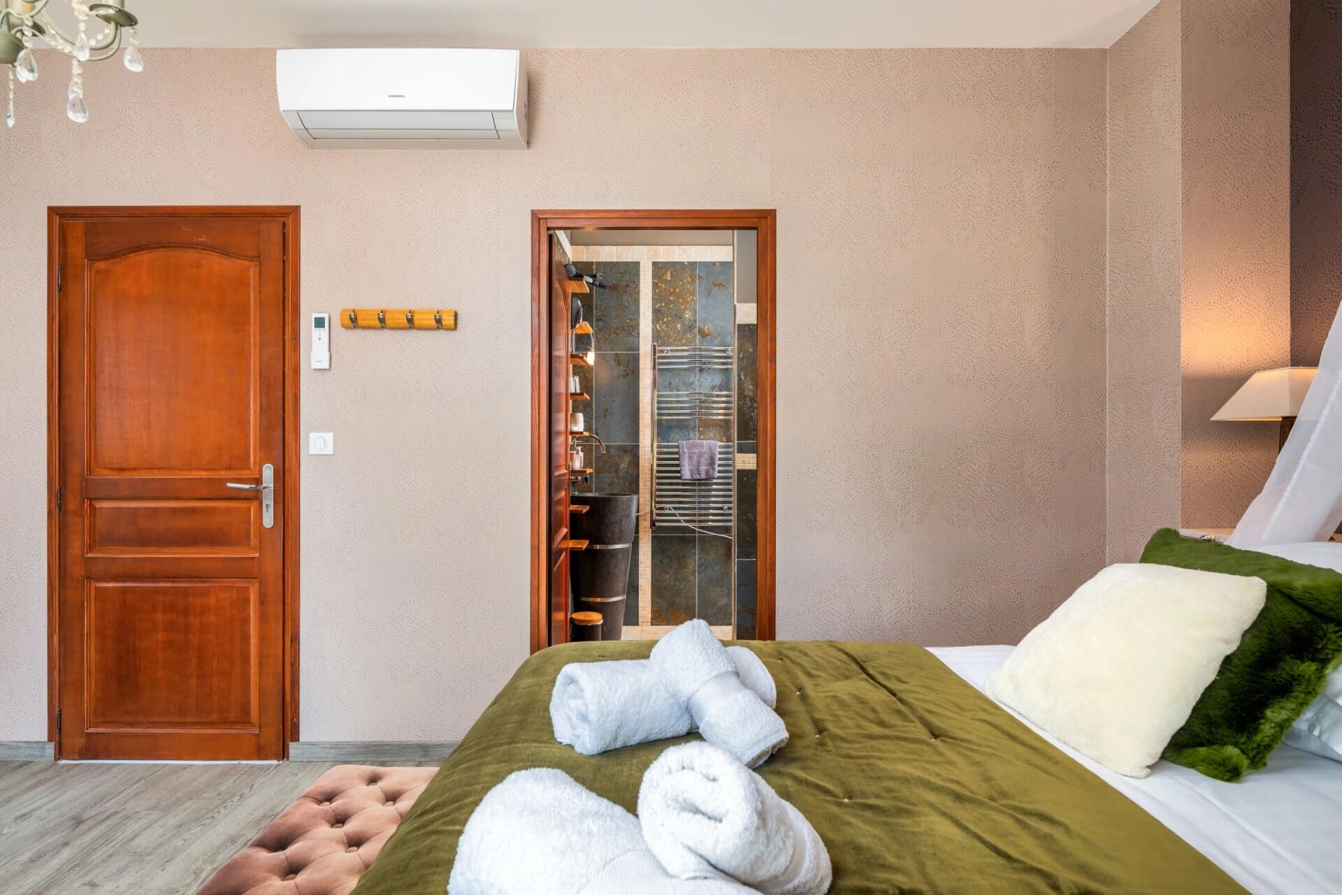 Habitación de hotel con puerta de madera, un minibár, cama con ropa de cama verde y almohadas blancas, y un pequeño banquillo tapizado.