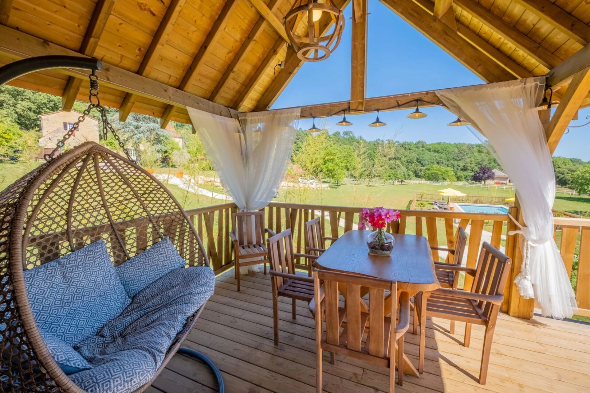Una terraza de madera con una silla colgante, una mesa con sillas y vistas a una piscina y follaje.