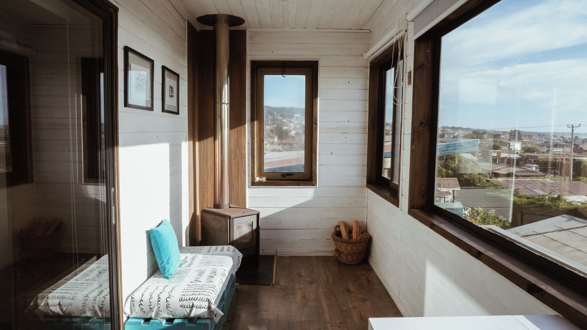 Una habitación pequeña y acogedora, con piso de madera, una cama con colcha estampada, una mesita y una ventana que ofrece vistas a un pueblo, además de cuadros en la pared.