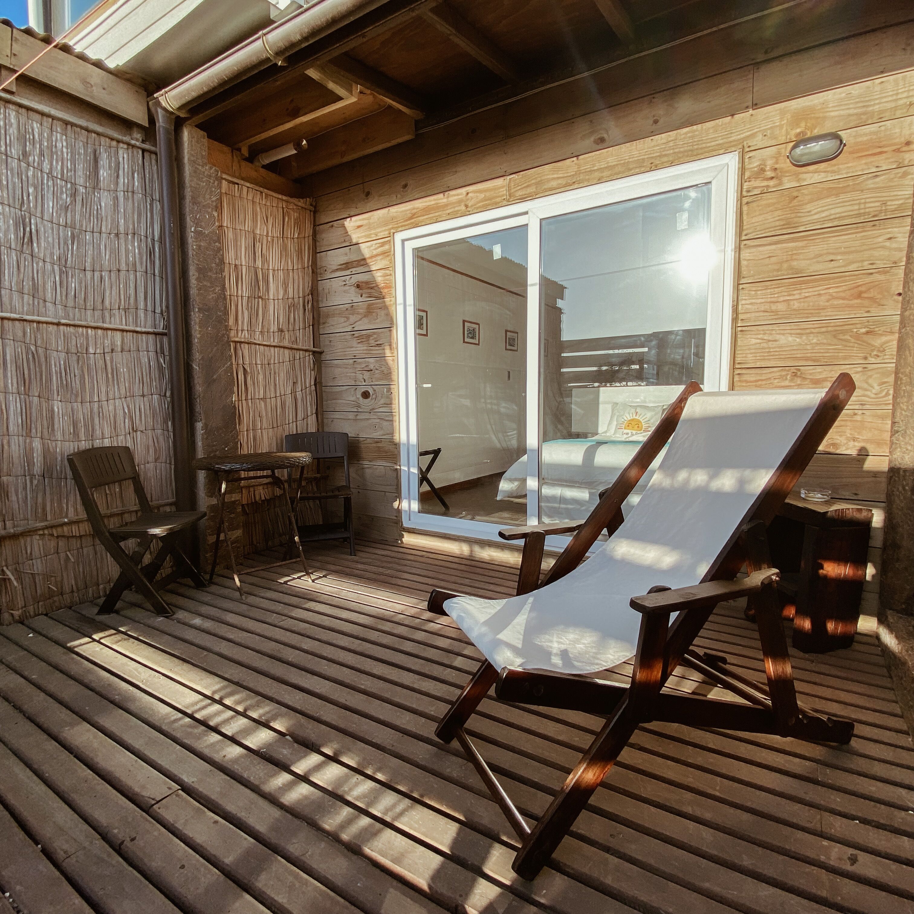 Terraza de madera con una silla y una mesa, puerta corrediza de vidrio que da a un dormitorio y pared de madera.