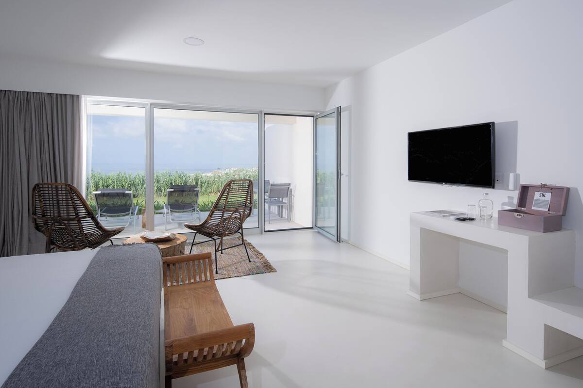 A modern hotel room with a large sliding glass door leading to a balcony, a flat-screen TV mounted on the wall, a wooden bench, and a small desk with a mirror and a pink box.