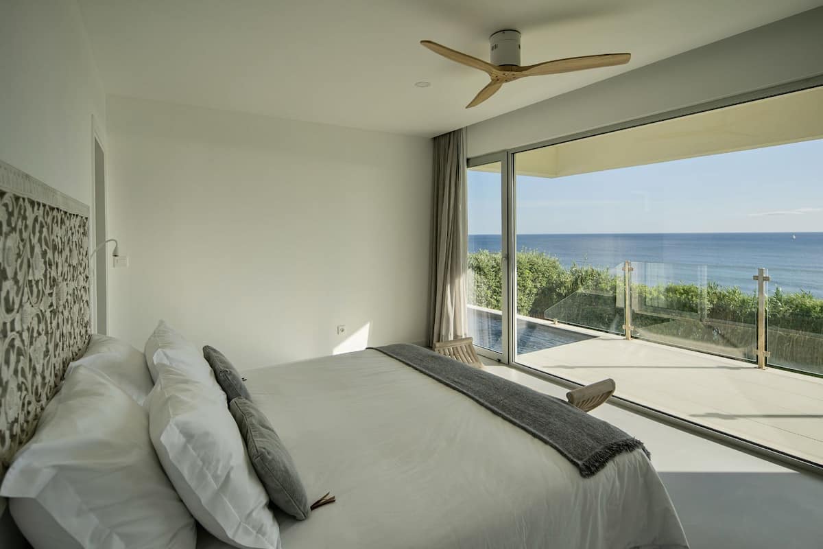 A bedroom with a large bed, a ceiling fan, and a balcony with a view of the sea.