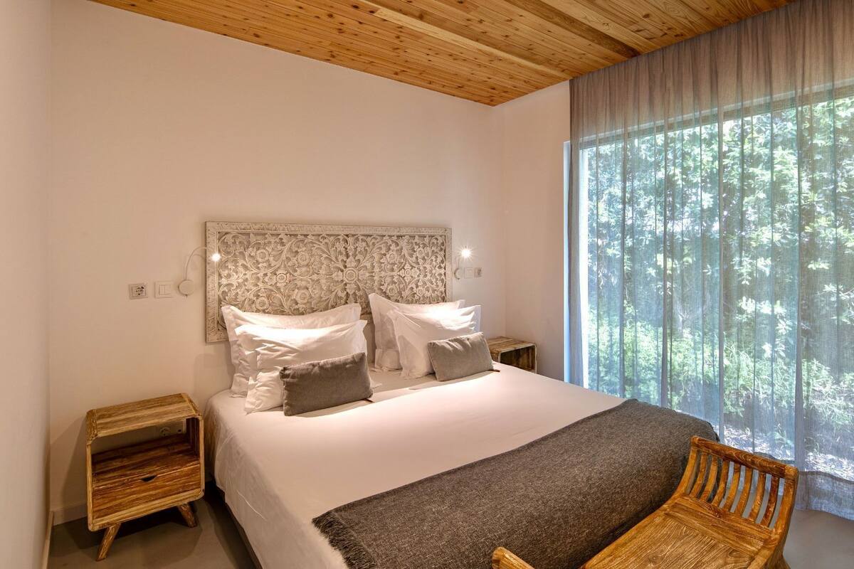 A bedroom with a large bed, a headboard with intricate carvings, bedside tables, a chair, and a large window with a view of trees.