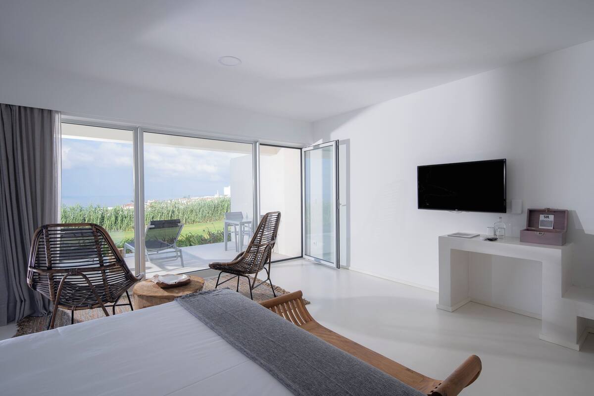 A modern hotel room with a large bed, two chairs, a small table, a TV, and a balcony with a view.