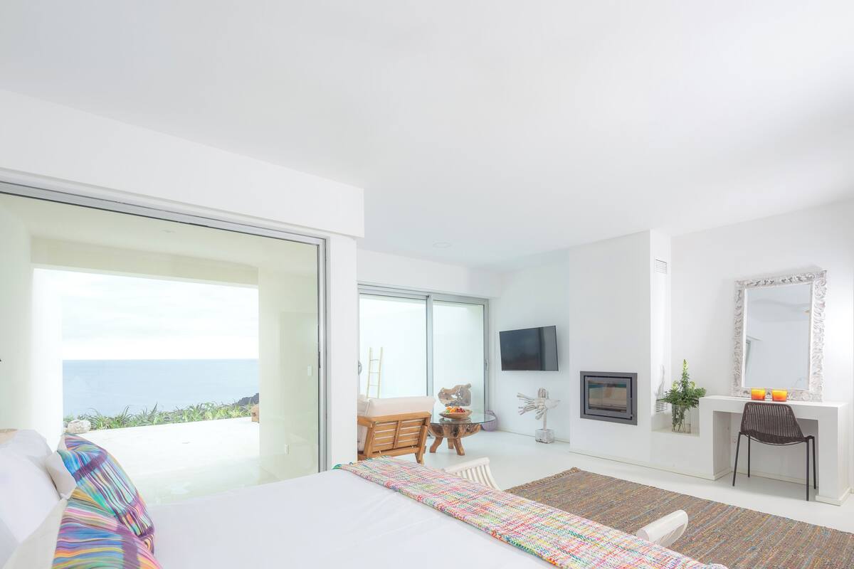 A modern bedroom with a large bed, a desk, a chair, a television, and a view of the ocean.