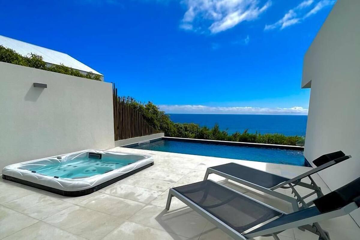 A rooftop pool area with a hot tub and lounge chairs, overlooking a clear blue sky and ocean.