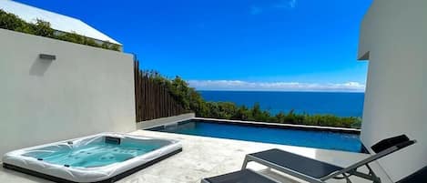 A rooftop pool area with a hot tub and lounge chairs, overlooking a clear blue sky and ocean.
