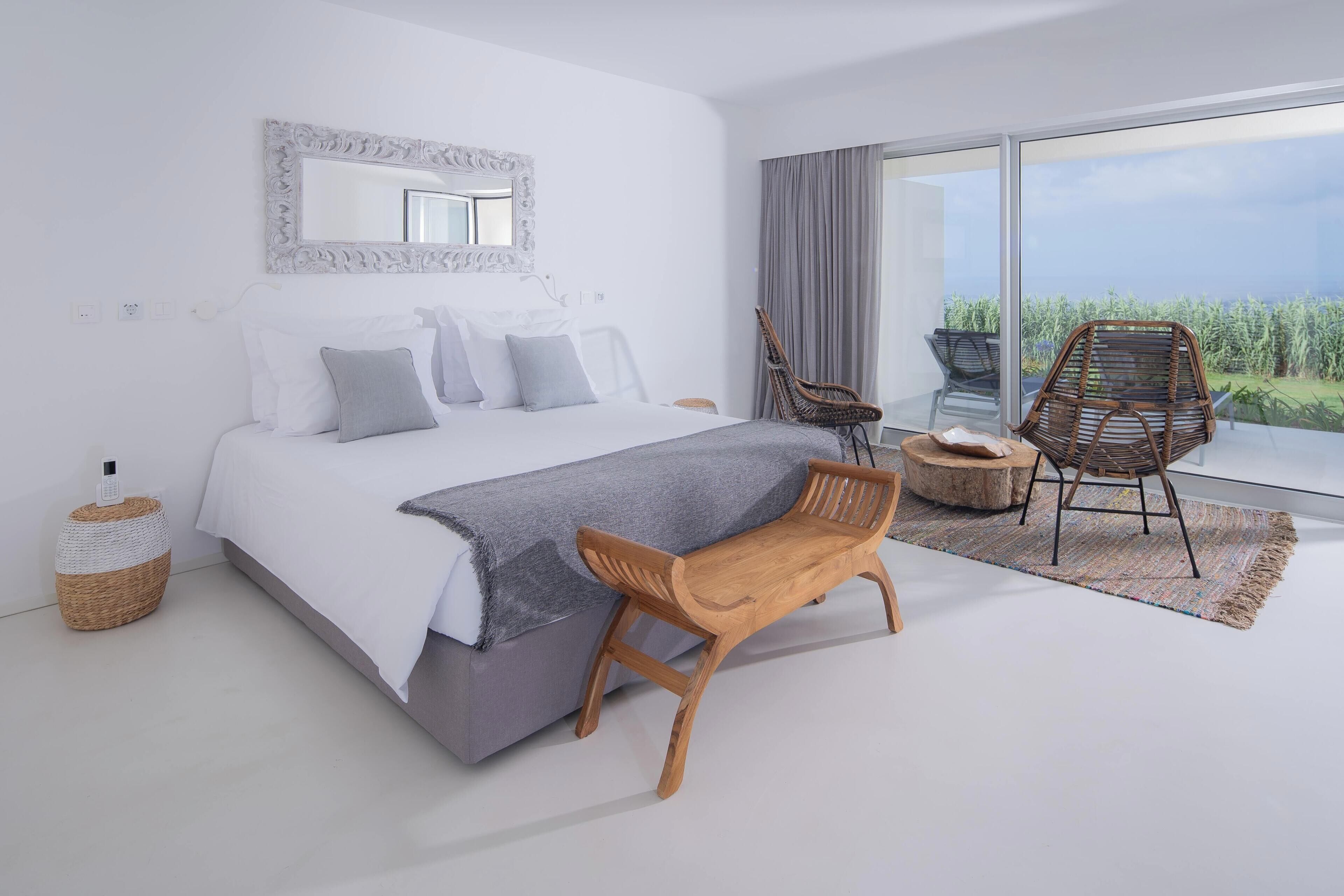 Um quarto moderno com uma cama grande, um banco de madeira, duas cadeiras de vime e vista para o campo através de amplas janelas.