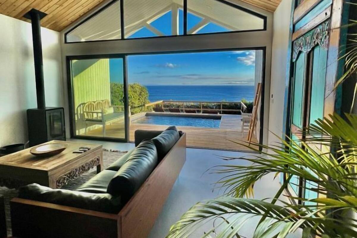 A modern living room with a large glass door leading to a pool area, wooden ceiling, and a view of the sea.