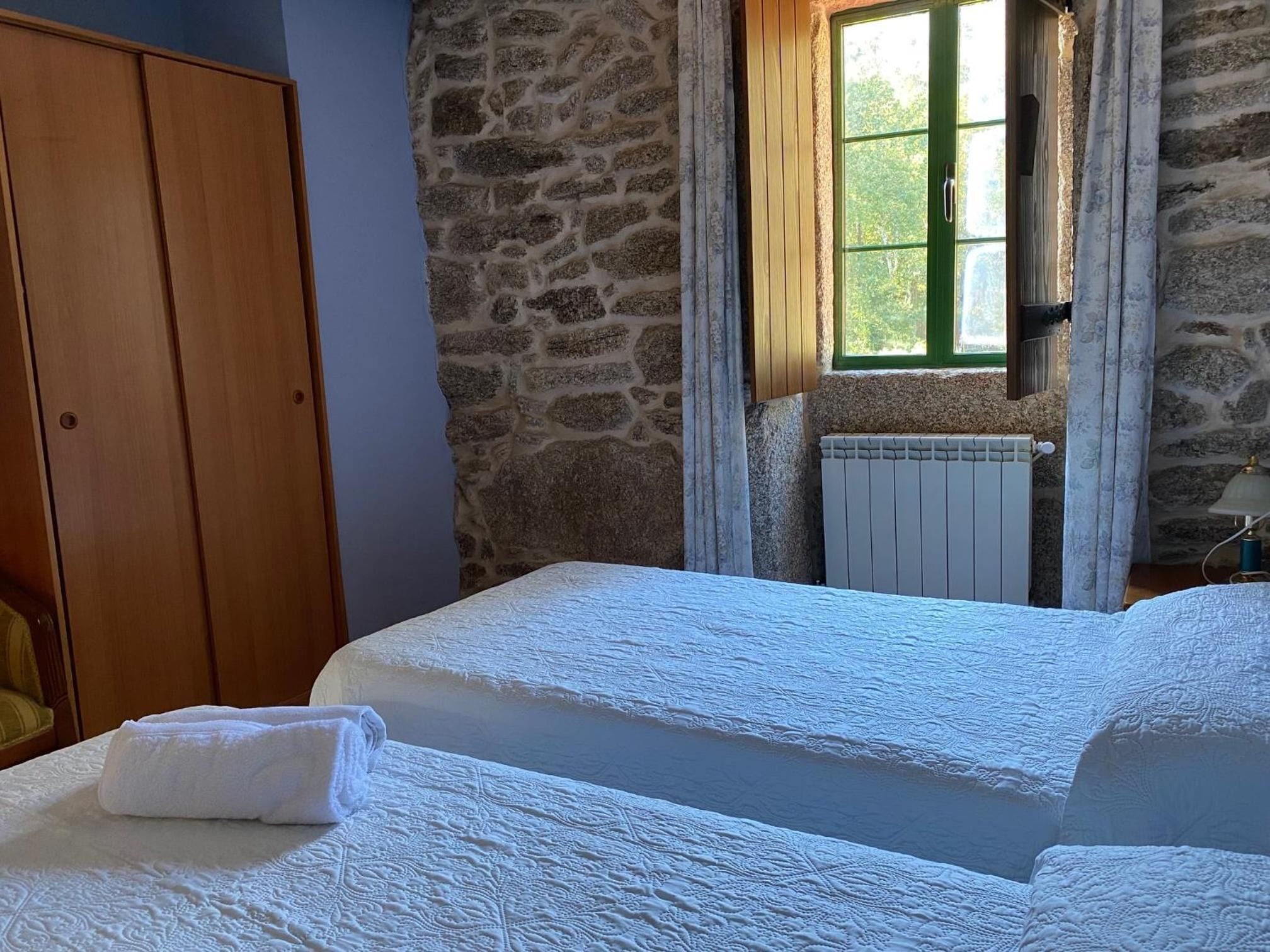 A bedroom with a stone wall, a wooden wardrobe, a bed with white bedding, and a window with curtains.
