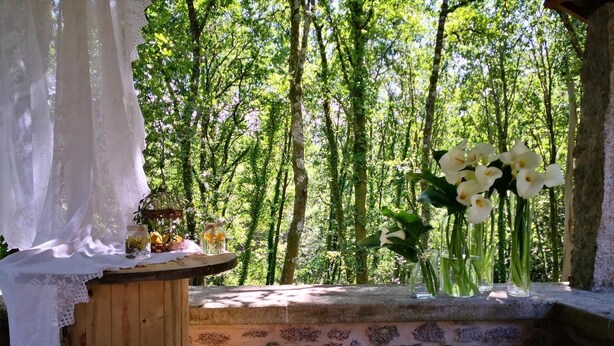 Eine rustikale Außenkulisse mit einem Holztisch, einer weißen Tischdecke mit Spitzenbordüre und einer Vase mit weißen Blumen.