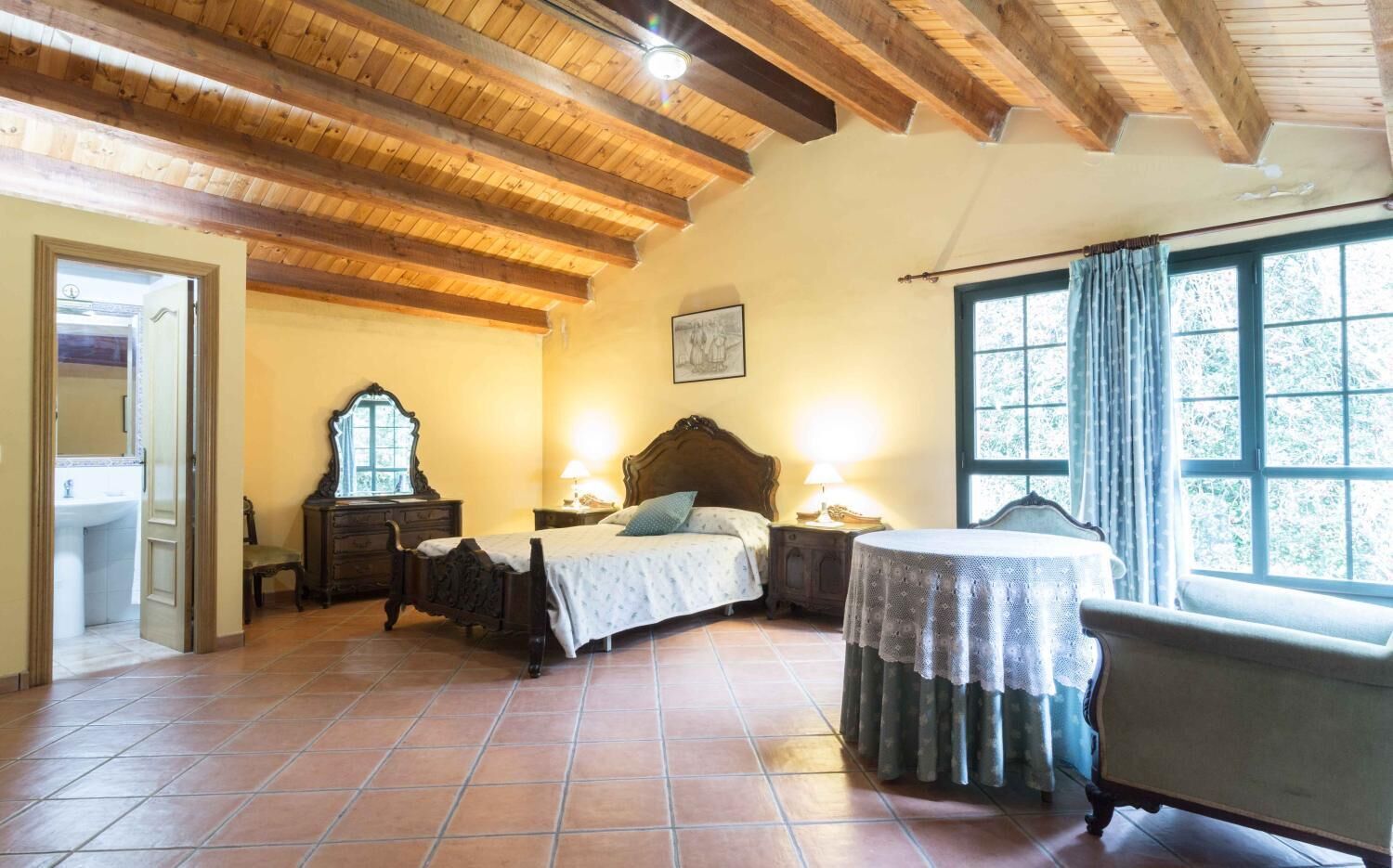 Un dormitorio con techo de madera, una cama con cabecera, una mesita de noche con lámpara, una silla y una ventana con cortinas.