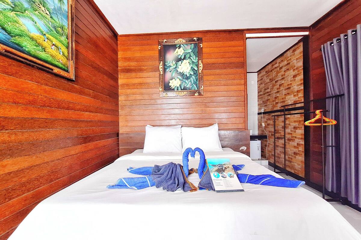 A hotel room with a wooden wall, a painting, a neatly made bed with towels, and a hanging coat rack.