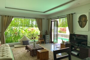 A living room with a sofa, a coffee table, a TV, and a view of greenery through large windows.