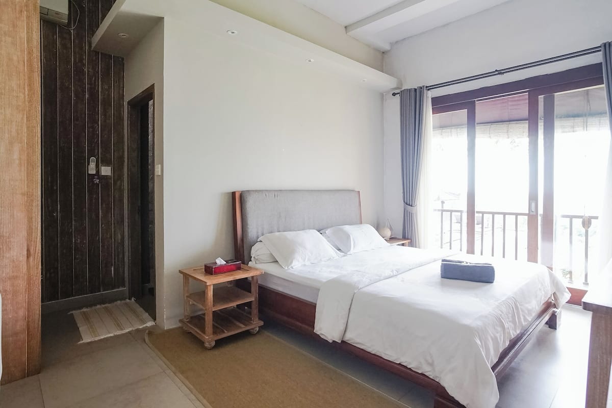 Ein Hotelzimmer mit einem großen Bett, einem Holfnachtschrank, einem kleinen Tisch und einem Balkon mit Vorhängen.