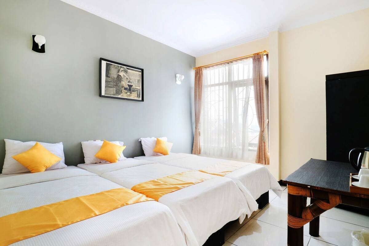 A hotel room with two beds, a framed picture on the wall, a window with curtains, and a small table with a kettle.