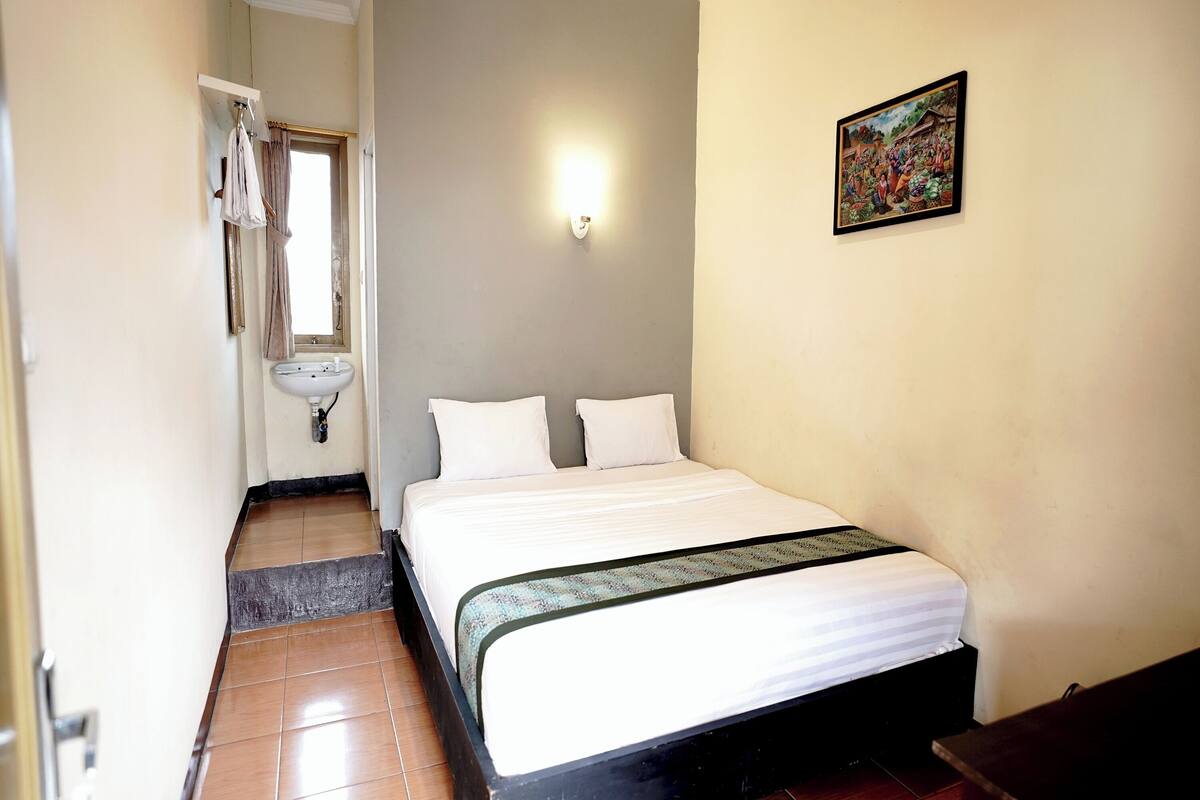 A bedroom with a bed, a window, a sink, and a painting on the wall.