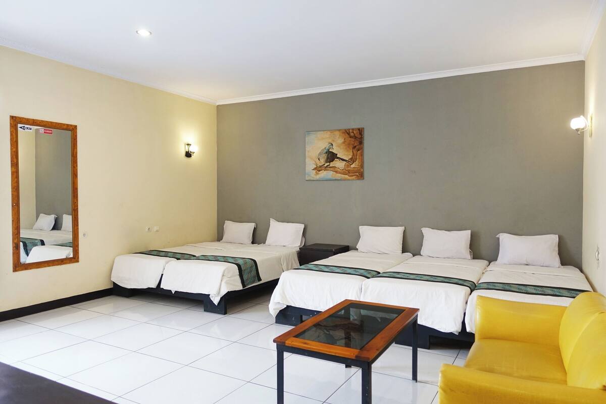 A hotel room with two single beds, a yellow armchair, a small table, a painting on the wall, and a mirror with a sign.