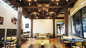 Restaurante com tetos altos, vigas aparentes e luminárias pendentes. Mesas e cadeiras dispostas para refeições.