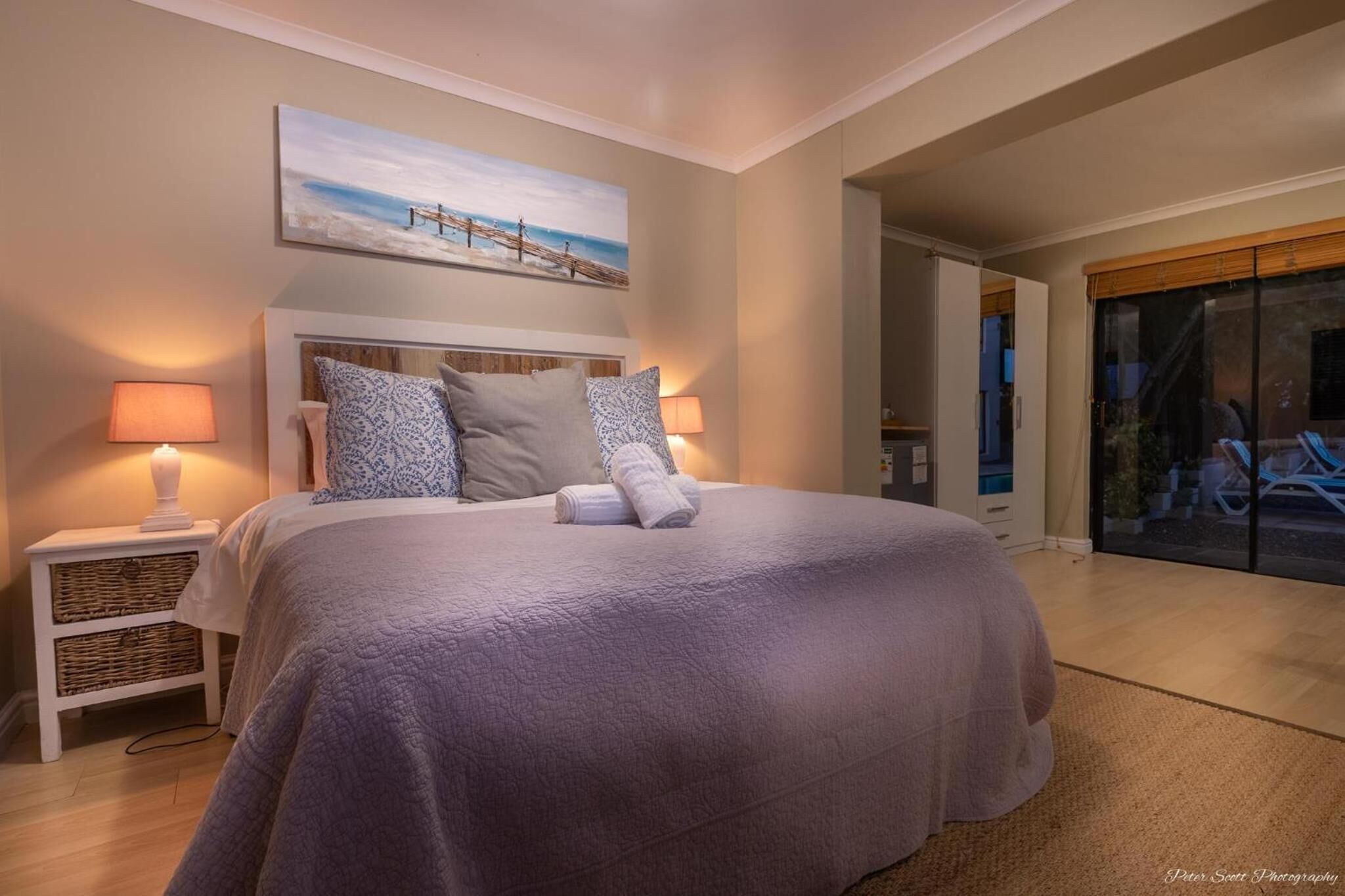 A neatly made bed with a headboard, two bedside lamps, and a painting above it.